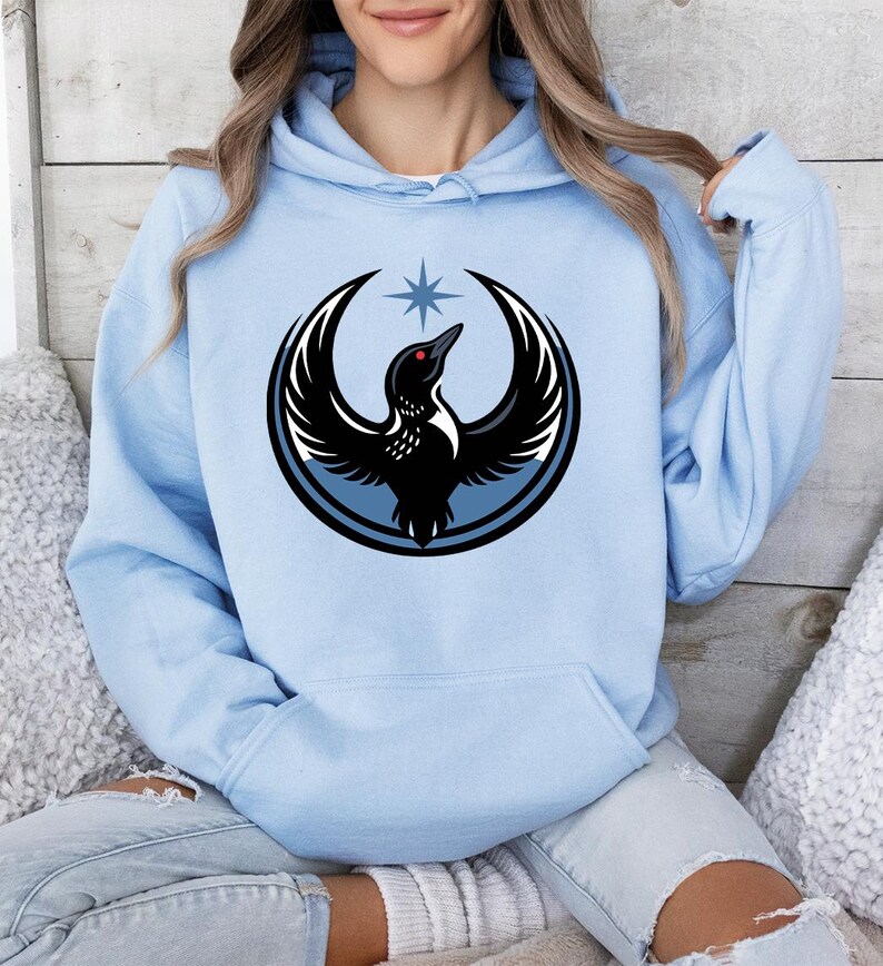 May include: Light blue hoodie featuring a circular graphic of a black bird with outstretched wings, a red eye, and a star above its head. The bird is set against a light blue background with a black outline. The hoodie has a front pocket and a drawstring hood.
