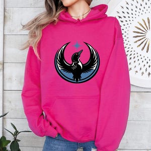 May include: A vibrant pink hoodie featuring a graphic of a bird with outstretched wings, a blue circle, and a star. The bird is black and white. The hoodie has a front pocket and a hood.