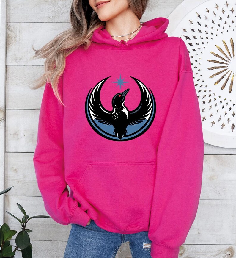 May include: A vibrant pink hoodie featuring a graphic of a bird with outstretched wings, a blue circle, and a star. The bird is black and white. The hoodie has a front pocket and a hood.