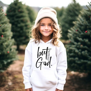 May include: A white hoodie featuring the words "but God" in black script. The hoodie also has the text "There was no way, But God made a way." The person in the image is wearing a beige beanie.