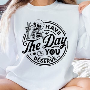 May include: White sweatshirt featuring a black graphic of a skeleton giving a peace sign. The text reads "I HAVE The Day YOU DESERVE" in a circular design. A casual, comfortable top.