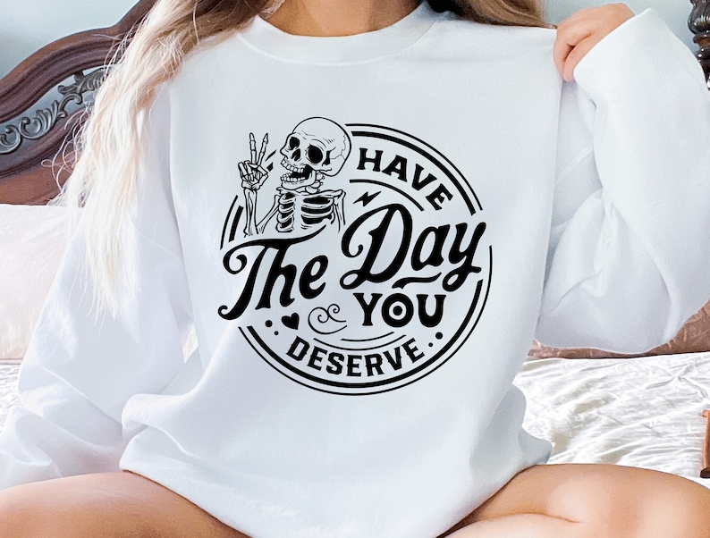 May include: White sweatshirt featuring a black graphic of a skeleton giving a peace sign. The text reads "I HAVE The Day YOU DESERVE" in a circular design. A casual, comfortable top.