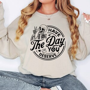 May include: Beige sweatshirt with a black graphic. The graphic features a skeleton giving a peace sign, with the words "Have The Day You Deserve" in a circular design. The sweatshirt has long sleeves and a crew neck.