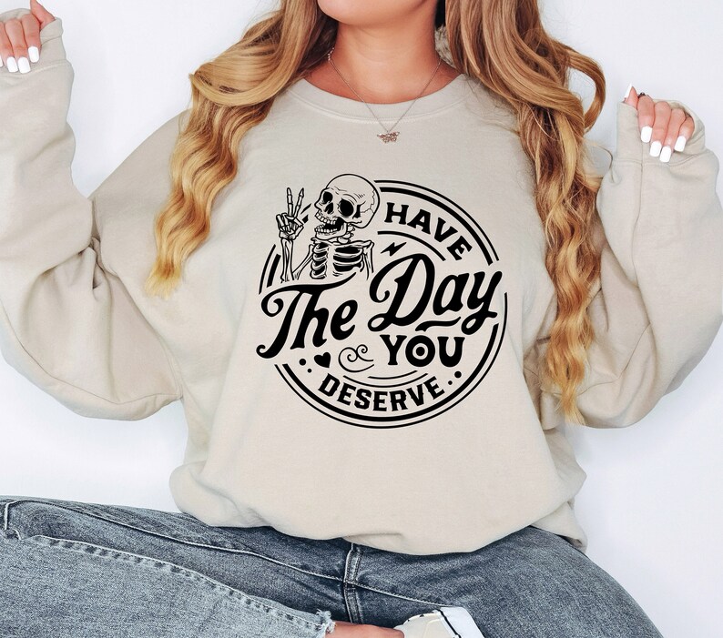May include: Beige sweatshirt with a black graphic. The graphic features a skeleton giving a peace sign, with the words "Have The Day You Deserve" in a circular design. The sweatshirt has long sleeves and a crew neck.