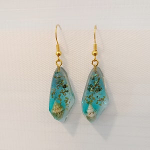 Könnte beinhalten: Ein Paar goldene Ohrringe mit blauen, tropfenförmigen Anhängern aus Harz. Die Anhänger enthalten goldenen Glitzer, kleine Muscheln und andere natürliche Elemente.
