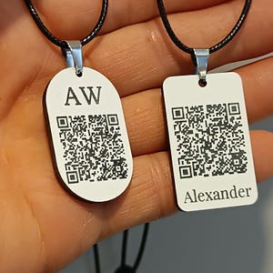 Könnte beinhalten: Zwei Halsketten mit Anhängern. Ein ovaler Anhänger mit "AW" und einem QR-Code, der andere rechteckig mit "Alexander" und einem QR-Code. Beide weißen Anhänger sind an schwarzen Kordeln mit silberfarbenen Metallbefestigungen befestigt.