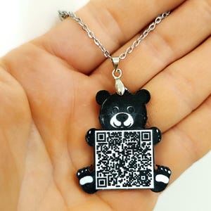 Könnte beinhalten: Eine schwarze Bären-Anhänger-Halskette mit einer silbernen Kette. Der Bär hat einen weißen Bauch und Pfoten, und ein QR-Code befindet sich in der Mitte. Die Halskette wird in einer Hand gehalten.