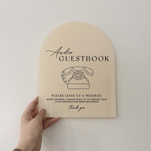 May include: A beige acrylic sign with rounded edges shaped like an arch. The sign has black text that reads "Audio Guestbook". A black outline of a rotary phone is below the text. The sign also has the text "Please leave us a message. When finished, please hang up to ensure that your message has been recorded. Thank you".
