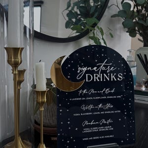 May include: A dark blue arched acrylic sign with white text and star graphics, featuring a gold crescent moon accent. The sign lists signature drinks including To the Moon and Back, Written in the Stars, and Moonbeam Martini, supported by a clear base.