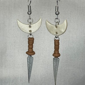 May include: A pair of silver earrings with a crescent moon charm and a silver dagger charm. The dagger charm has a brown handle.