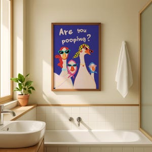May include: A framed art print featuring a blue background with the text "Are you pooping?" and illustrations of three geese wearing sunglasses and scarves. The artwork is displayed in a bathroom setting, with a white towel and a potted plant visible.