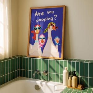 May include: A framed art print featuring three geese wearing sunglasses and scarves, set against a blue background. The text "Are you pooping?" is displayed in white. The artwork is in a bathroom setting, with green tiles and bath accessories.