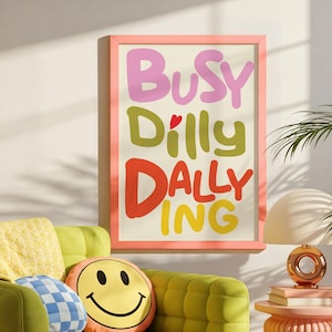 May include: A framed art print with the words "Busy Dilly Dallying" in colorful, playful lettering. The pink-framed artwork hangs on a white wall above a green sofa with a smiley face pillow.