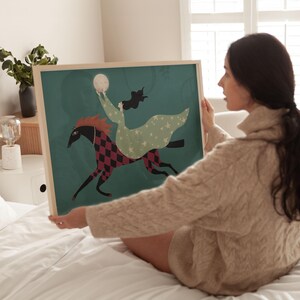 May include: A framed art print featuring a whimsical illustration of a woman riding a horse. The horse has a black head and a red and black diamond pattern on its body. The woman wears a green dress with white stars and holds a moon.