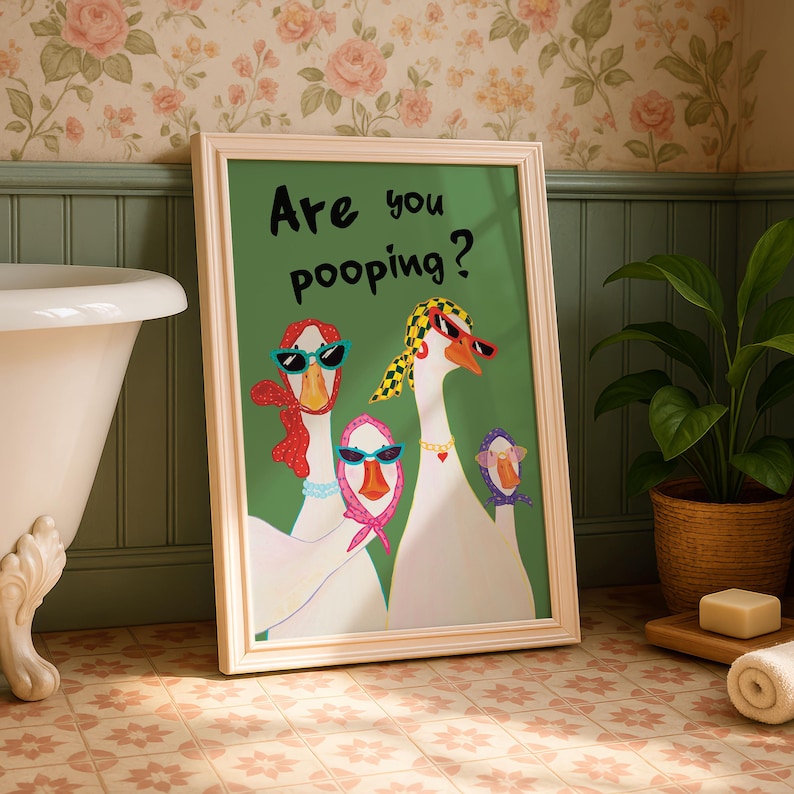 May include: A framed art print featuring three geese wearing sunglasses and scarves, with the text "Are you pooping?" on a green background. The artwork is in a white frame, leaning against a wall with a floral wallpaper.