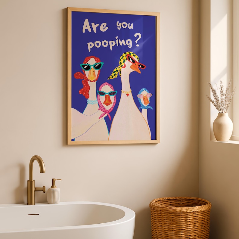 May include: A framed art print featuring a whimsical illustration of three geese wearing sunglasses and scarves against a blue background. The text "Are you pooping?" is displayed at the top. The artwork is in a light wood frame, adding a touch of humor to the bathroom decor.