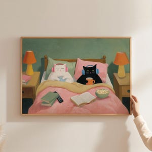 May include: A framed art print showing two cats in bed, each wearing headphones. One is white, the other black, both are holding mugs. The bed has a pink comforter, pillows, and a bowl of popcorn. Two bedside tables with lamps flank the bed.