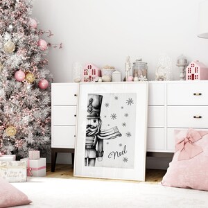 May include: A black and white illustration of a nutcracker soldier with a red scarf and a white background. The word "Noel" is written in cursive below the nutcracker. There are snowflakes scattered around the nutcracker.