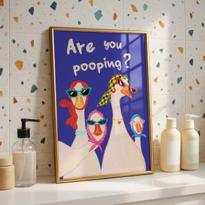 May include: A framed art print featuring a blue background with the text "Are you pooping?" and illustrations of geese wearing sunglasses and scarves. The frame is gold.
