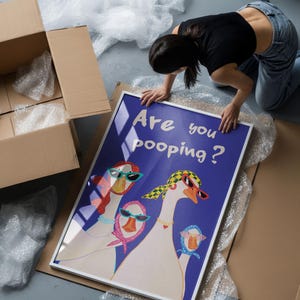 May include: A framed art print featuring a whimsical illustration of geese wearing sunglasses and scarves, set against a blue background. The text "Are you pooping?" is displayed above the geese. The print is being handled near cardboard boxes and bubble wrap.