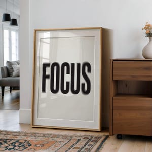 Può includere: Un poster incorniciato con la parola "FOCUS" stampata in nero su sfondo bianco. Il poster è appeso a una parete bianca in una stanza con pavimento in legno.
