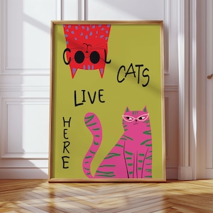 以下が含まれることがあります： ライムグリーンの背景に「COOL CATS LIVE HERE」の文字が入った額入りのアートプリント。サングラスをかけた赤い猫とメガネをかけたピンクの猫のイラストが描かれています。フレームはライトブラウンです。