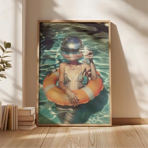 May include: A framed art print featuring a person in a pool wearing a disco ball head and a swimsuit, holding a cocktail. They are floating in a pool with an orange and white life preserver. The artwork has a vintage, retro aesthetic.