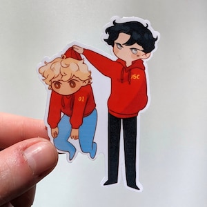 以下が含まれることがあります： 2人の漫画のキャラクターが描かれたステッカー。 「JSC」と書かれた赤いパーカーを着た一人は、別の赤いパーカーのフードを持っています。 2番目のキャラクターは青いズボンを履いており、パーカーには番号「01」が書かれています。 白い背景。