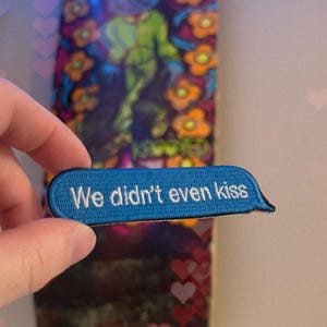 Może przedstawiać: Niebieska, haftowana naszywka w kształcie dymka z tekstem "We didn't even kiss". Naszywka jest trzymana przed deską do skateboardu z kolorowym wzorem przedstawiającym postać z kreskówki i elementy kwiatowe.