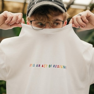 May include: White crew neck sweatshirt with the text "JOY IS AN ACT OF RESISTANCE" in rainbow-colored embroidery. The sweatshirt is being held up by a person wearing a gray baseball cap and glasses.