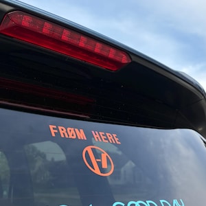 May include: A black car's rear window features orange and blue decals. The top decal reads "FROM HERE" above an orange logo. Below, the blue text says "HAVE A GOOD DAY". The sky is visible in the background.