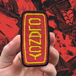May include: A rectangular patch with the word "OZORO" embroidered in yellow on a red background. The patch has a black border and is held in a hand. The background is a collage of red and black papers.