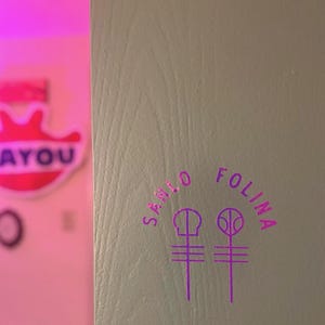 May include: A close-up of a light gray surface with the words "SANLO FOLINA" in a purple, stylized font. Below the text are two purple symbols. The background is a blurred pink and white sign that says "AYOU".