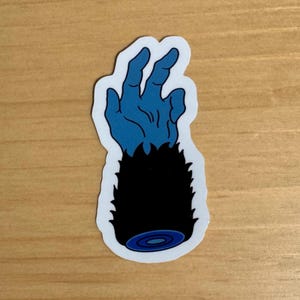 Può includere: Una mano blu con pelliccia nera che si alza, con un cerchio blu e bianco alla fine del braccio.