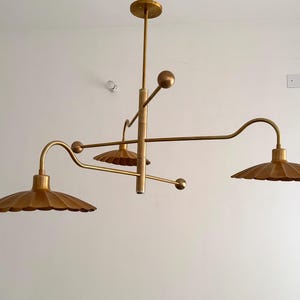May include: A gold-toned, contemporary chandelier featuring three lampshades. The light fitting has a central vertical support with horizontal and angled arms, each with a scalloped lampshade. Spherical accents are placed along the arms.