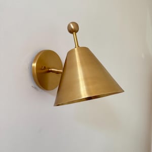 May include: A brass wall sconce featuring a conical shade and a circular backplate. The light fitting has a small spherical detail at the top. It is mounted on a white wall. The fixture is made of metal.