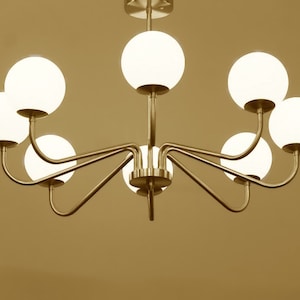 K&ouml;nnte beinhalten: Ein goldfarbener Kronleuchter mit acht wei&szlig;en, kugelf&ouml;rmigen Gl&uuml;hbirnen. Der Kronleuchter hat ein schlichtes, modernes Design mit geschwungenen Armen.