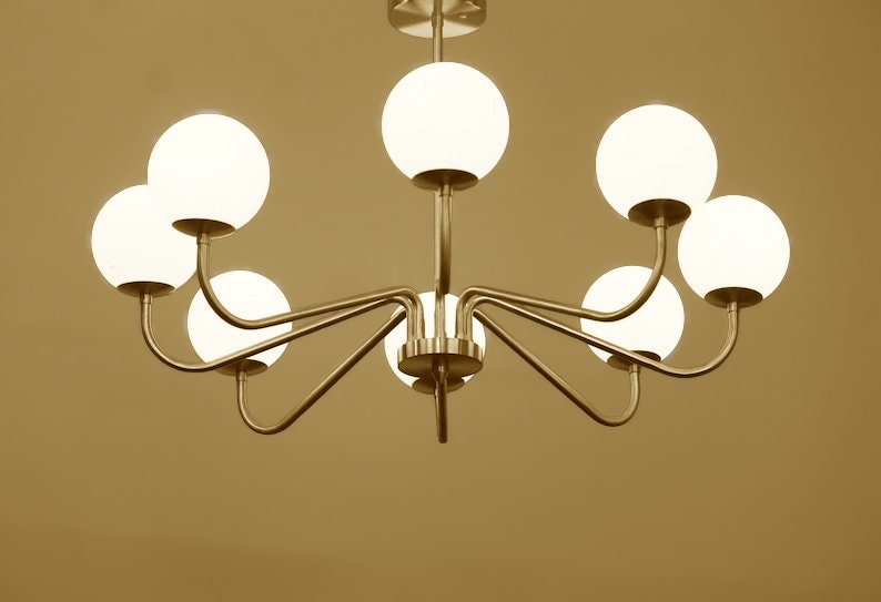 K&ouml;nnte beinhalten: Ein goldfarbener Kronleuchter mit acht wei&szlig;en, kugelf&ouml;rmigen Gl&uuml;hbirnen. Der Kronleuchter hat ein schlichtes, modernes Design mit geschwungenen Armen.