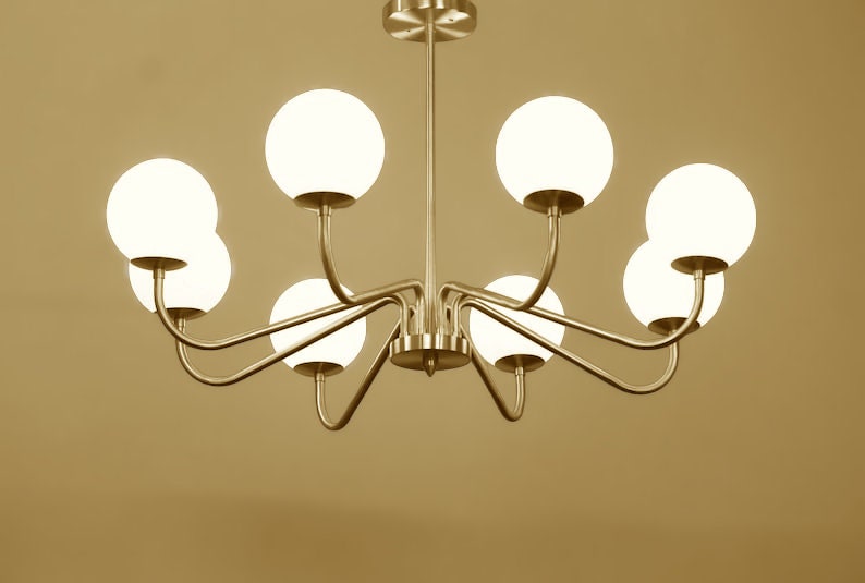 K&ouml;nnte beinhalten: Ein goldfarbener Kronleuchter mit acht wei&szlig;en, kugelf&ouml;rmigen Gl&uuml;hbirnen. Der Kronleuchter hat ein modernes Design mit geschwungenen Armen.