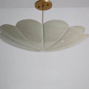 Può includere: Una moderna plafoniera a forma di petalo. La lampada ha un paralume color crema e uno stelo e un supporto a soffitto in metallo dorato. L'apparecchio è progettato per essere montato a soffitto e fornisce illuminazione ambientale.