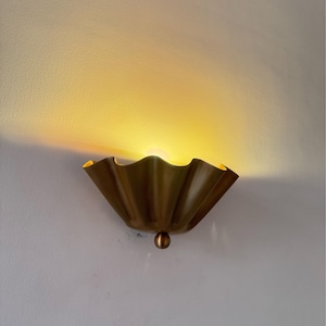 May include: A gold-colored wall sconce with a scalloped, shell-like design. The light fixture has a small, round accent at the bottom and emits a warm, yellow glow. The sconce is mounted on a white wall.