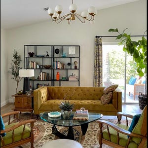 K&ouml;nnte beinhalten: Ein Wohnzimmer mit einem goldenen Samtsofa, zwei gr&uuml;nen Sesseln, einem Glas-Couchtisch, einem wei&szlig;en Hocker und einem schwarzen Metall-Regal. Der Raum hat einen gemusterten Teppich, eine wei&szlig;e Decke und einen goldenen Kronleuchter mit wei&szlig;en Kugeln.