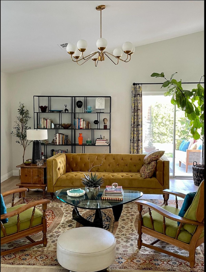 K&ouml;nnte beinhalten: Ein Wohnzimmer mit einem goldenen Samtsofa, zwei gr&uuml;nen Sesseln, einem Glas-Couchtisch, einem wei&szlig;en Hocker und einem schwarzen Metall-Regal. Der Raum hat einen gemusterten Teppich, eine wei&szlig;e Decke und einen goldenen Kronleuchter mit wei&szlig;en Kugeln.