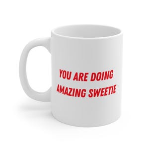 Könnte beinhalten: Weiße Keramik-Kaffeetasse mit dem roten Text "YOU ARE DOING AMAZING SWEETIE". Die Tasse hat einen gebogenen Henkel und eine glänzende Oberfläche. Der Text ist fett und ohne Serifen.