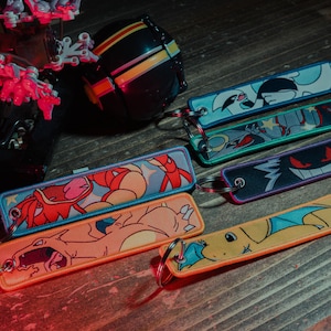May include: Rectangular keychains with various cartoon character designs. The keychains feature characters in orange, blue, purple, and green. Each keychain has a metal ring for attachment.