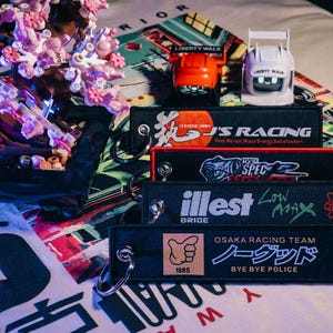 May include: An assortment of black keychains with embroidered text and graphics, including "J'S RACING", "NYC SPEC", "illest", and "OSAKA RACING TEAM". Also, two toy cars, one orange and one white, are visible. The keychains have metal rings.
