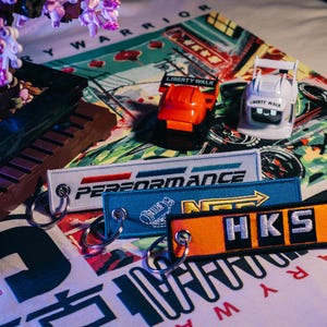 May include: An assortment of automotive-themed accessories, including keychains with "PERFORMANCE" and "HKS" text, and miniature car models. The items are arranged on a patterned surface with Japanese text and floral accents.