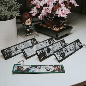 May include: Five black and white anime character keychains with black and white printed designs. The keychains are on a white surface. One keychain has a green and black design with the text "My" printed on it.