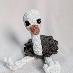 May include: A handmade crochet ostrich doll. The ostrich has a white head and neck, a brown beak, and black eyes. The body is dark gray, and the legs and feet are white. The doll is sitting on a white surface.