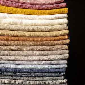 May include: A stack of folded linen fabric in various colors, including pink, mustard yellow, cream, and shades of brown, gray, and blue. The fabrics are neatly arranged, showcasing their textures and color variations. The stack is on a wooden surface.